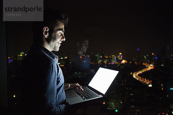 Junger Geschäftsmann tippt nachts vor dem Fenster eines Bürohochhauses am Laptop  Shanghai  China