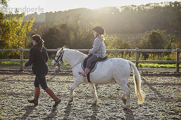 Ausbilderin führt Mädchen auf weißem Pony reitend auf dem Reitplatz
