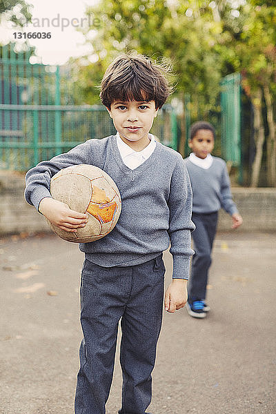 Porträt eines Grundschülers  der einen Fussball auf dem Spielplatz hält