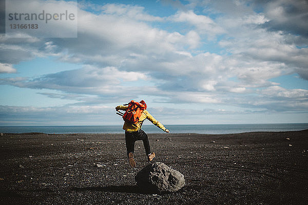Rückansicht eines mittelgroßen Mannes  der über einen Felsbrocken auf vulkanische Landschaft springt  Island