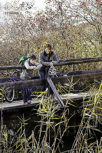 Mutter und Sohn mit Fahrrädern stehen auf einem Holzsteg und schauen auf den Fluss