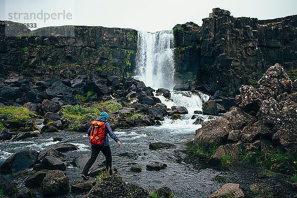 Rückansicht einer mittelgroßen erwachsenen Frau mit Rucksack  die über Felsen im Fluss vor einem Wasserfall tritt  Island