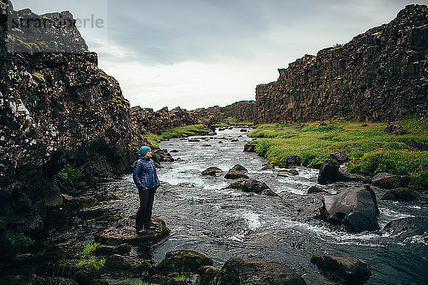 Mittelgroße erwachsene Frau steht auf einem Felsen im Flussbett und schaut weg  Island