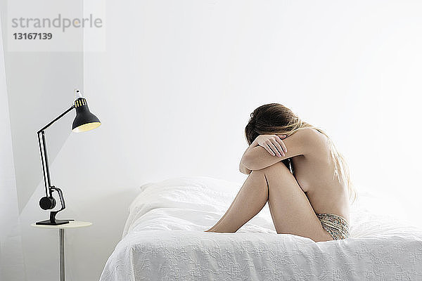 Junge Frau sitzt auf dem Bett  umarmt die Knie und versteckt ihr Gesicht