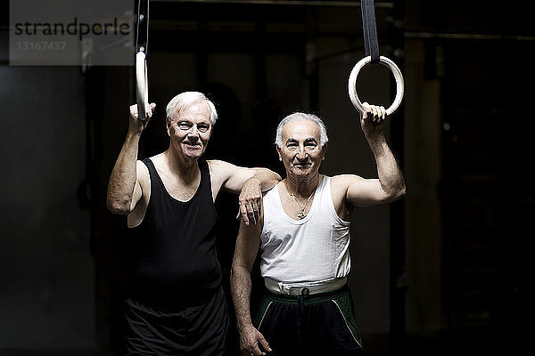 Porträt von zwei älteren Männern  die in dunkler Turnhalle Turnringe halten