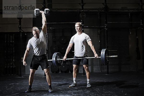 Zwei junge Männer beim Gewichtheben mit Hantel und Langhantel in dunkler Turnhalle