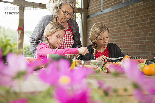 Zwei Schwestern und eine Großmutter am Küchentisch lernen frisches Obst zu schneiden