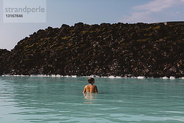 Rückansicht einer mittelgroßen erwachsenen Frau  die mit der Taille tief im Wasser steckt  Heiße Quellen in der Blauen Lagune  Island