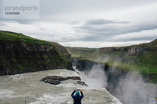 Hochwinkel-Rückansicht einer mittelgroßen erwachsenen Frau  die ein Smartphone benutzt  um einen Wasserfall zu fotografieren  Island