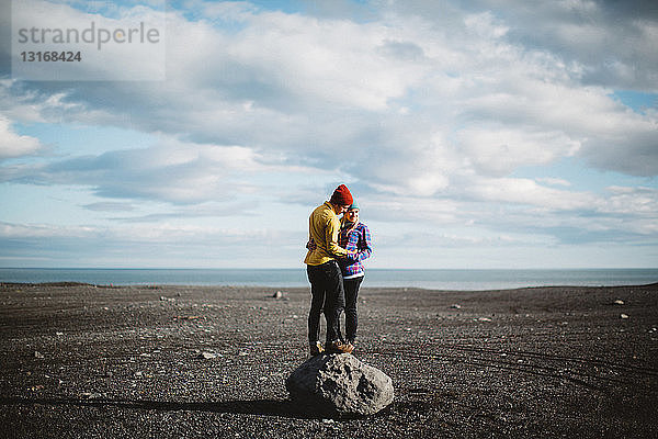 Mittelgroßes erwachsenes Paar auf einem Felsblock stehend  von Angesicht zu Angesicht in vulkanischer Landschaft umarmt  Island