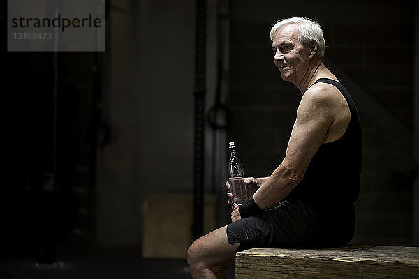 Älterer Mann sitzt mit Wasserflasche in dunkler Turnhalle