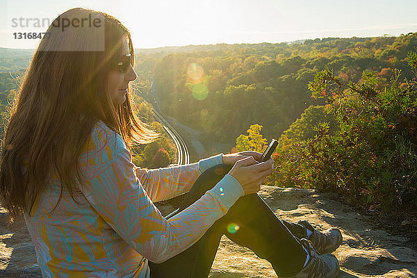 Mittelgroße erwachsene Frau sitzt auf einem erhöhten Felsen und benutzt ein Mobiltelefon