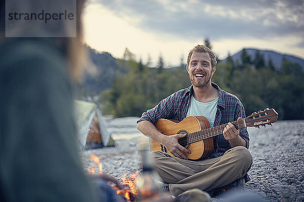 Frontansicht eines jungen Mannes  der am Lagerfeuer sitzt und Gitarre spielt