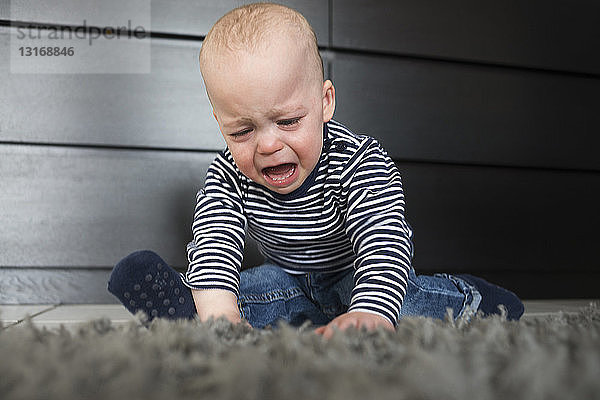 Weinender kleiner Junge sitzt auf einem Teppich im Wohnzimmer