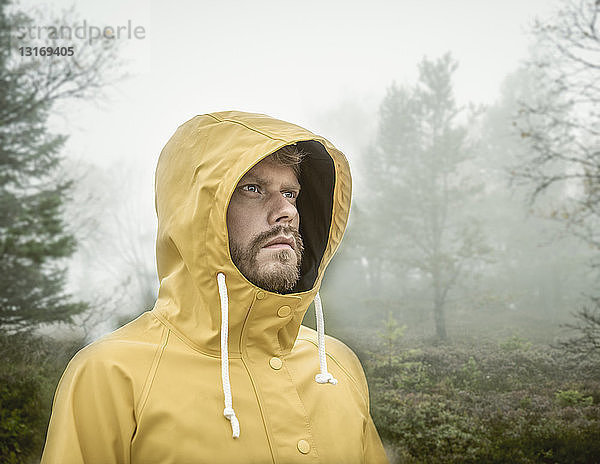 Bärtiger junger Mann in nebligem Wald mit gelbem Regenmantel mit Kapuze schaut weg