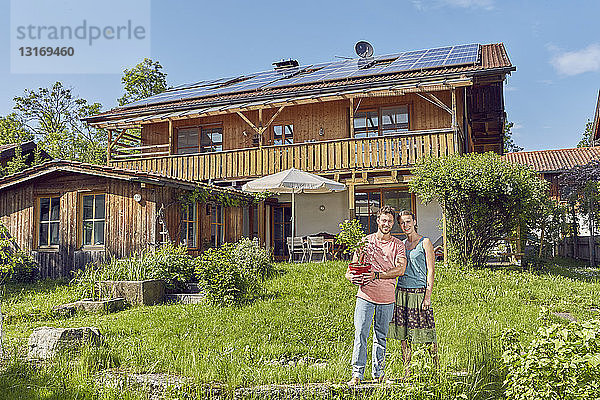 Porträt eines jungen Paares mit einer Topfpflanze in der Hand  stehend vor einem Haus mit Sonnenkollektoren