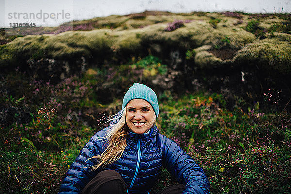 Frontansicht einer mittelgroßen erwachsenen Frau  die mit einer Strickmütze vor dem Hügel kauert und lächelnd in die Kamera schaut  Island