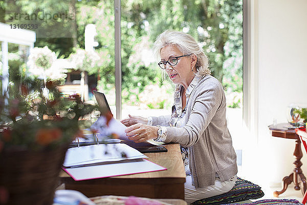 Ältere Frau am Tisch sitzend  Papierkram durchsehen  Laptop benutzen