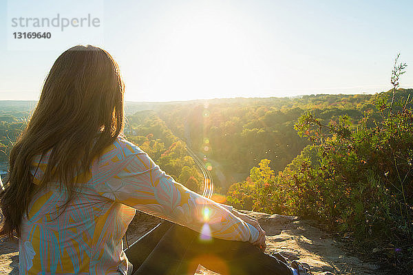 Mittelgroße erwachsene Frau sitzt auf einem erhöhten Felsen und betrachtet den Sonnenaufgang