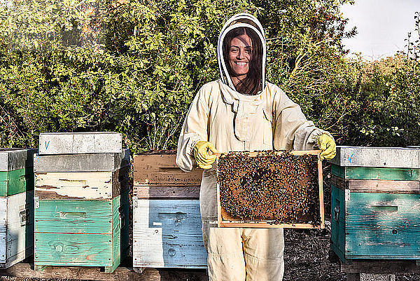 Porträt einer Frau in Imkerkleidung  die einen mit Bienen bestückten Bienenstock hält