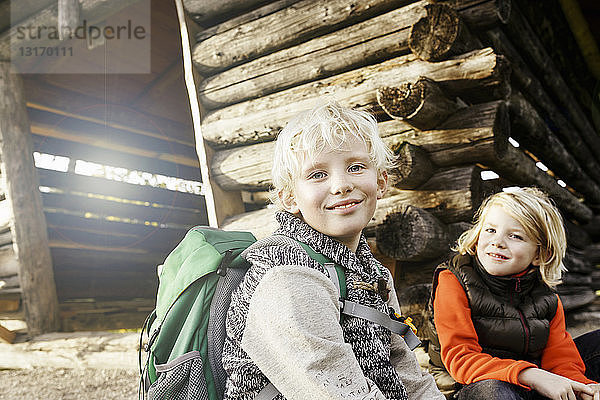 Blondhaarige Geschwister sitzen vor der Blockhütte und schauen lächelnd in die Kamera