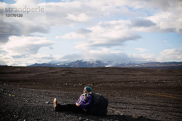 Seitenansicht einer mittelgroßen erwachsenen Frau  die an einen Felsbrocken gelehnt auf einer vulkanischen Landschaft sitzt und wegschaut  Island