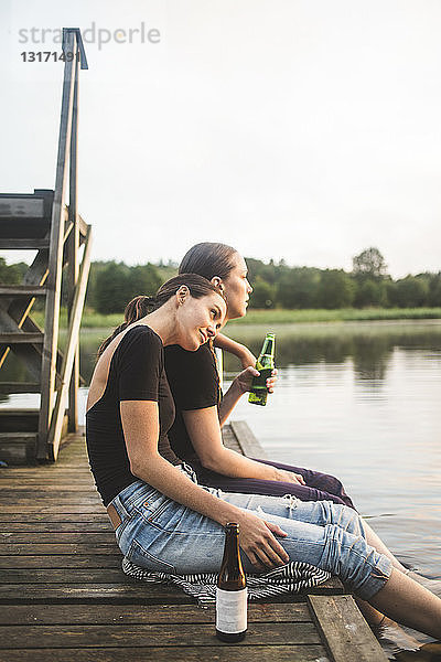 Seitenansicht von Freundinnen  die auf den See schauen  während sie bei Sonnenuntergang auf dem Steg sitzen