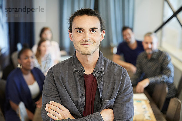 Porträt eines selbstbewussten Geschäftsmannes im Stehen  während Kollegen im Hintergrund im Kreativbüro arbeiten