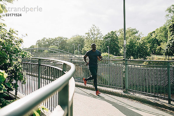 Mittlerer erwachsener Sportler in voller Länge joggt bei Sonnenschein auf einer Fußgängerbrücke in der Stadt
