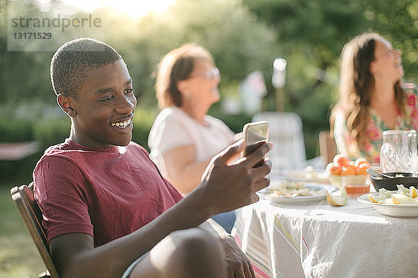Junge lächelt beim Benutzen eines Mobiltelefons im Garten während einer Gartenparty