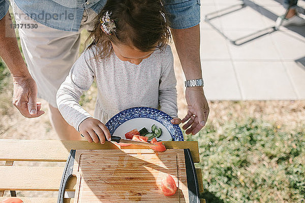 Mädchen legt Tomatenscheiben mit dem Messer in einen Teller  während sie während der Gartenparty beim Großvater am Tisch steht