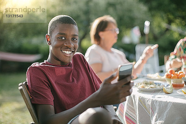 Porträt eines lächelnden Jungen  der während einer Gartenparty im Garten ein Mobiltelefon benutzt