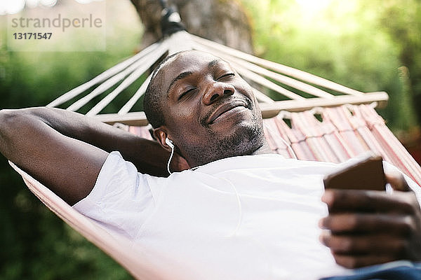 Lächelnder Mann mit geschlossenen Augen genießt Musik über Mobiltelefon  während er sich auf der Hängematte entspannt