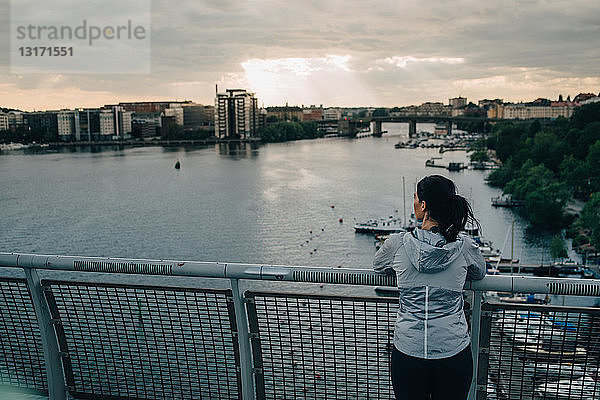 Rückansicht einer Athletin  die bei Sonnenuntergang auf einer Fußgängerbrücke über dem Meer steht und auf die Stadt schaut