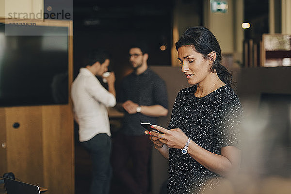 Textnachrichten von Geschäftsfrauen auf Smartphones mit Kollegen im Hintergrund im Büro