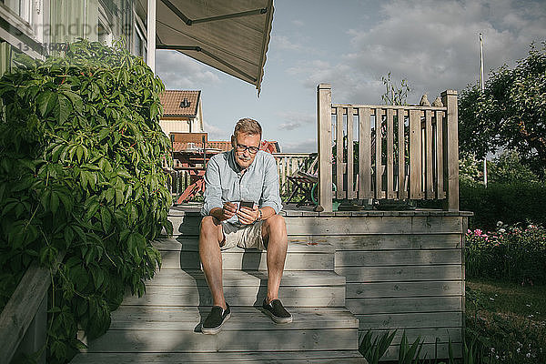Ein älterer Mann in voller Länge benutzt ein Mobiltelefon  während er auf einer Treppe auf der Veranda sitzt