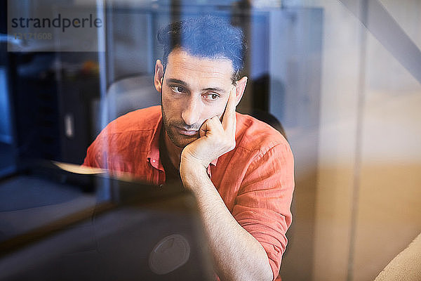 Nachdenklicher Geschäftsmann schaut durch Glas  während er im Büro am Computerschreibtisch sitzt