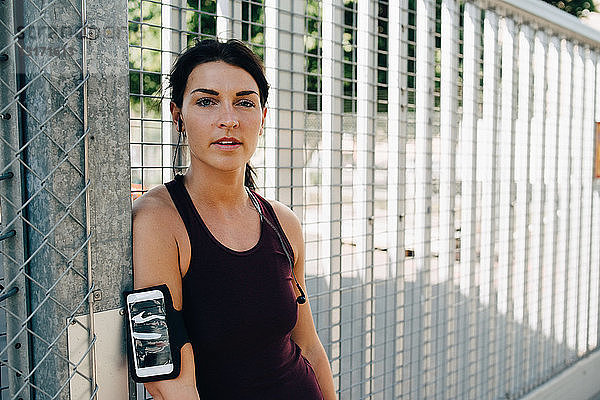 Porträt einer selbstbewussten Sportlerin mit Smartphone und In-Ear-Kopfhörern  die sich an der Brücke an ein Geländer lehnt