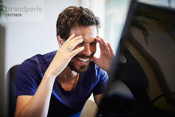 Fröhlicher Geschäftsmann schaut auf den Computermonitor  während er im Kreativbüro sitzt