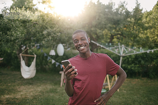 Porträt eines lächelnden Jungen mit Handy in der Hand  der während einer Gartenparty im Hinterhof steht