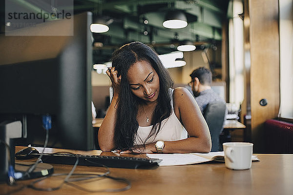 Geschäftsfrau mit Lesebuch mit der Hand im Haar am Schreibtisch im Büro