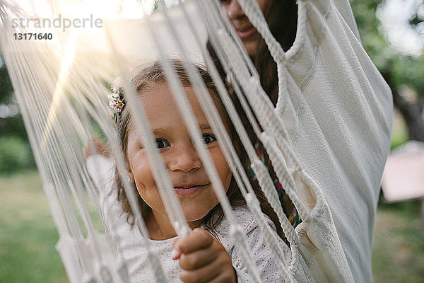 Porträt eines lächelnden Mädchens  das durch Seile gesehen wird  während es mit seiner Mutter auf einer Hängematte im Hinterhof schwingt