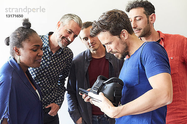 Geschäftsmann hält Virtual-Reality-Simulator in der Hand  während er Kollegen im Kreativbüro sein Handy zeigt