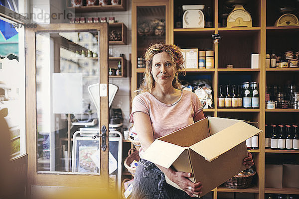 Porträt einer selbstbewussten Verkäuferin  die im Feinkostladen einen Karton gegen ein Regal trägt