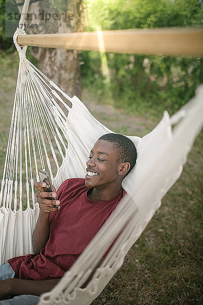 Lächelnder Junge benutzt Mobiltelefon  während er sich auf einer Hängematte im Hinterhof entspannt