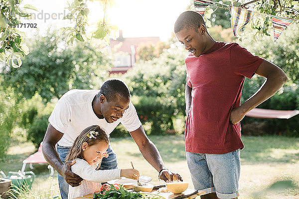 Junge sieht Vater an  der seiner Schwester bei der Zubereitung von Essen am Tisch während einer Gartenparty hilft