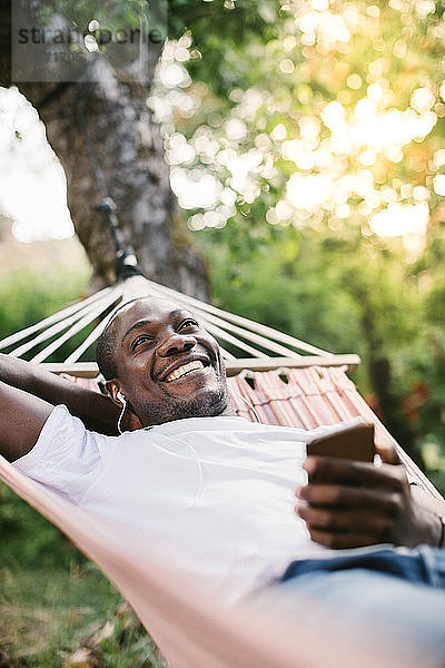 Lächelnder mittelgroßer Mann  der über sein Mobiltelefon Musik hört  während er sich auf einer Hängematte im Hinterhof entspannt