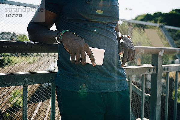 Mittelteil eines Sportlers  der ein Mobiltelefon hält  während er sich an einem sonnigen Tag an ein Geländer lehnt