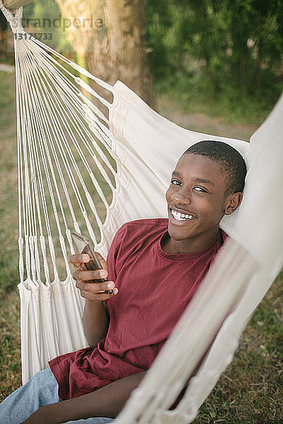 Porträt eines lächelnden Jungen  der ein Mobiltelefon benutzt  während er auf einer Hängematte im Hinterhof liegt