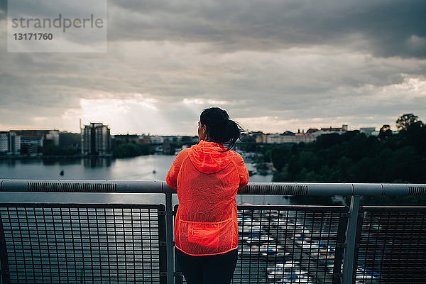 Rückansicht einer Sportlerin im Regenmantel  die bei Sonnenuntergang auf einer Fußgängerbrücke über dem Meer steht und auf die Stadt schaut